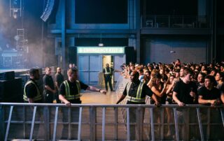 A concert stage view showing a security barricade and glowing exit sign before a packed audience.