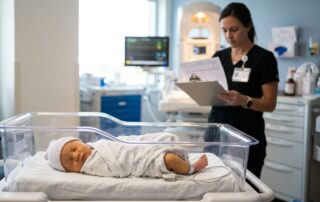A newborn rests in a clear bassinet while a clinician reviews bilirubin results.