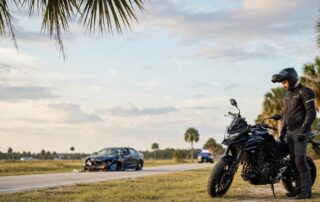Helmeted rider beside a motorcycle near a damaged sedan on a Florida roadside.