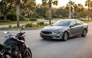 A motorcycle waits beside a scuffed car turning across its lane at a palm-lined intersection.